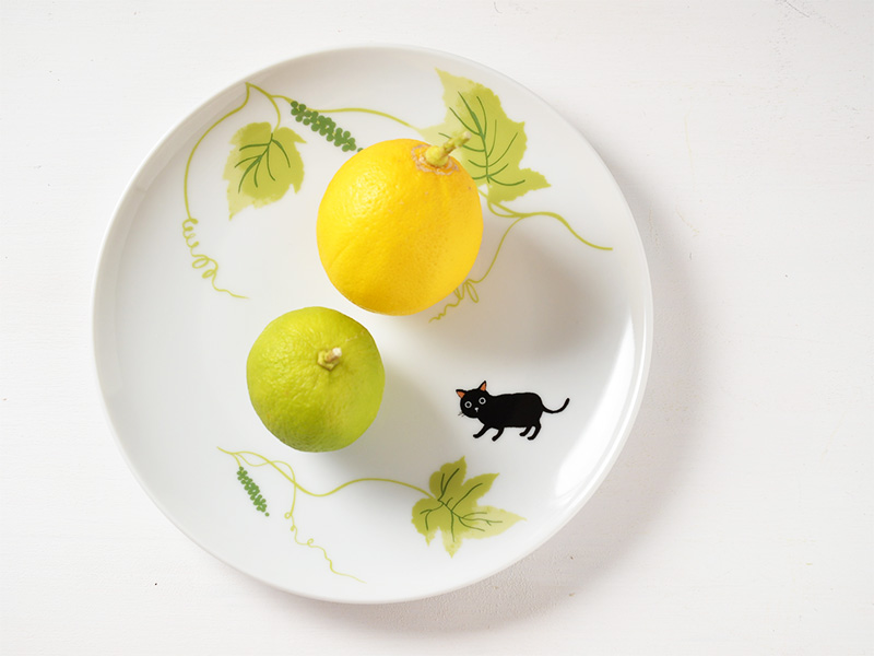 雑貨デザイナーシンジカトウさんが白い21cm位の丸い陶磁器のお皿に黒猫と緑色の葉っぱを描いたココシリーズの商品画像です。