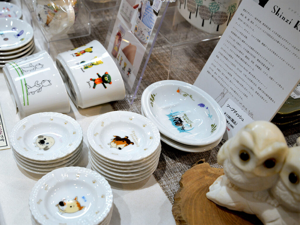 雑貨デザイナーシンジカトウさんデザインの犬のイラストの小皿や猫のイラストのソープディッシュ、くまのイラストの陶磁器製のスタンドのお店で販売している様子の画像です。