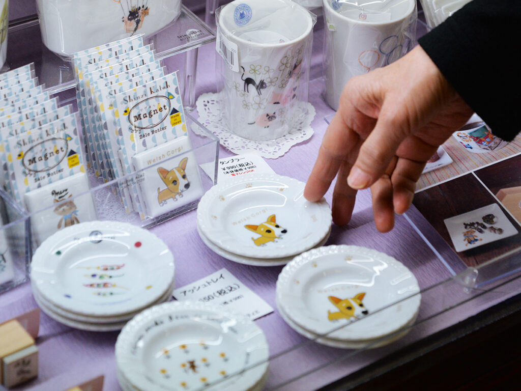 川島パーキングエリアの岐阜おみやげ川島店で販売されている雑貨デザイナーシンジカトウさんがデザインしたコーギーのイラストが描かれた小皿やマグネットタイルの陳列画像です。