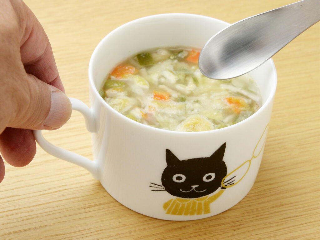 雑貨デザイナーシンジカトウさんがゼルポティエの為に描いたメガネを掛けた可愛い猫のイラストが美濃焼の白いマグカップに描かれたマグカップにスープを注いだ様子のイメージ画像です。