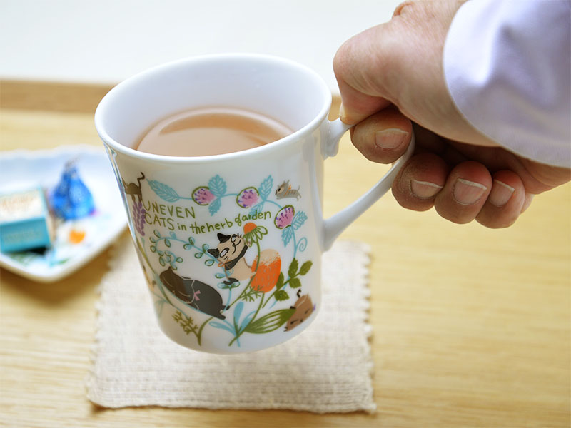 雑貨デザイナーシンジカトウさんデザインの可愛い猫のイラストのシリーズ「アニーブンキャッツシリーズ」の白さに拘った白磁の生地に猫達が草原の中でくつろぐ様子が描かれたマグカップを手に取ってトレーの上に置く様子の画像です。