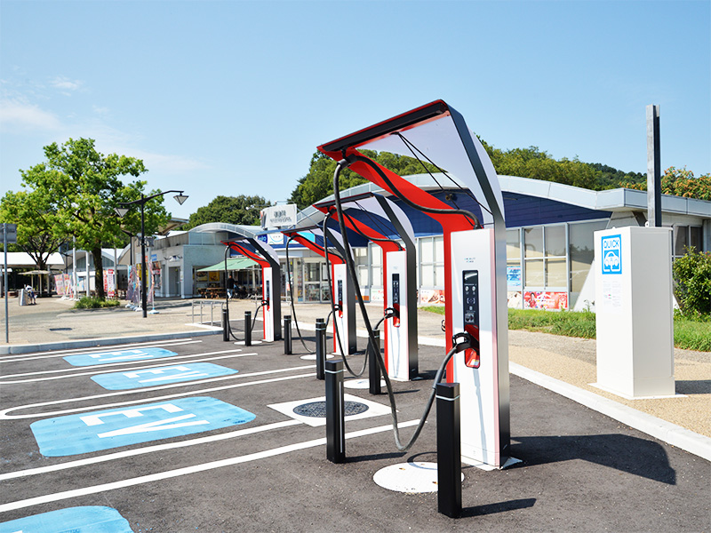 サービスエリアの駐車場にある、赤と白の電気自動車（EV）用充電スタンドが複数並んでいる様子。