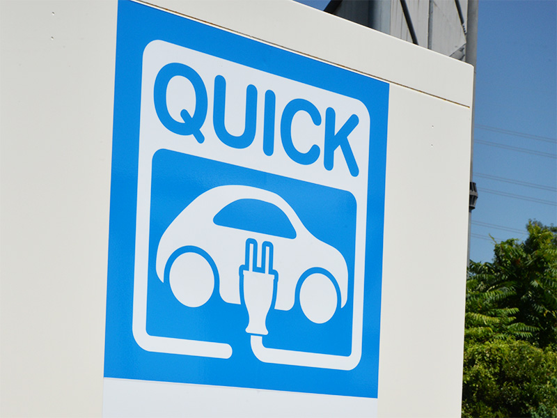 「QUICK」と書かれた青い文字の看板。電気自動車のイラストと充電プラグのマークが描かれており、会員や非会員向けの利用案内が日本語で書かれています。