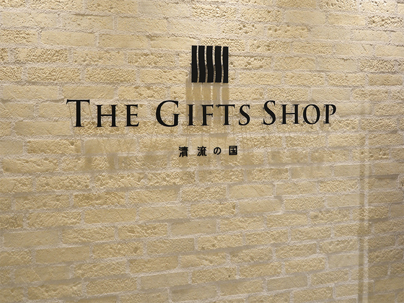 レンガ調の壁に黒色の立体文字で「The GIFTS SHOP」と中央に大きく書かれ、その下に少し小さいフォントで「清流の国」と印字されている看板。上部には黒い縦線が並んだシンボルマークがある。