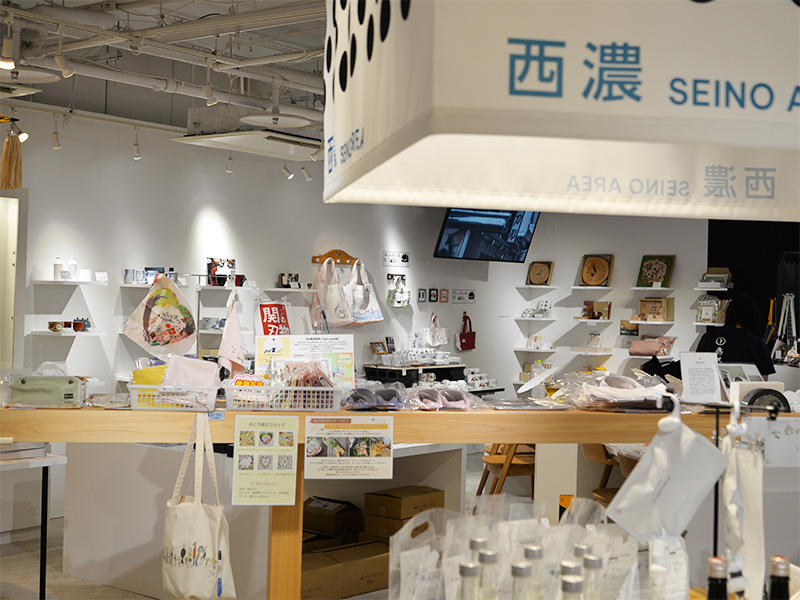 ギフトショップの店内を見下ろすようなアングル。天井からは「西濃 SEINO AREA」と書かれた看板が吊り下げられている。店内には様々な商品が並び、奥の壁には棚に商品がディスプレイされている様子が見える。手前には商品の詳細を紹介するPOP広告のようなものが見える。