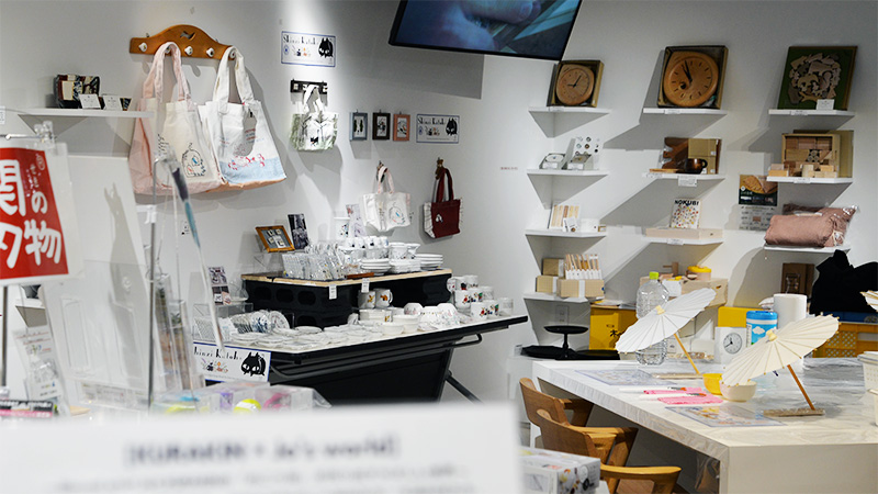 明るい店内、白壁に木製ハンガーに掛けられた猫のイラスト入りトートバッグ、壁に飾られた額縁、中央のテーブルに並べられた様々な猫や花柄デザインの白い美濃焼の食器類、奥の白い棚に飾られた木製時計や雑貨、手前に和傘が置かれたテーブルと椅子。全体的に温かみがあり、可愛らしい雑貨が豊富に並んでいるギフトショップの様子。