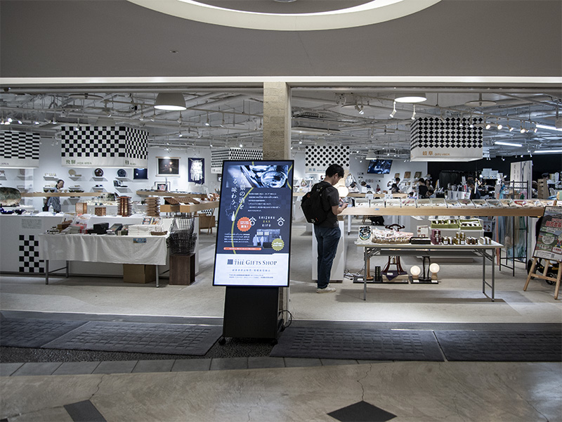 JR岐阜駅直結のアクティブG2階にあるギフトショップ「The GIFTS SHOP」の入り口付近。店内は明るく、様々な商品が並んでおり、奥にはお客さんの姿が見える。手前にはイベント告知のデジタルサイネージが立っている。