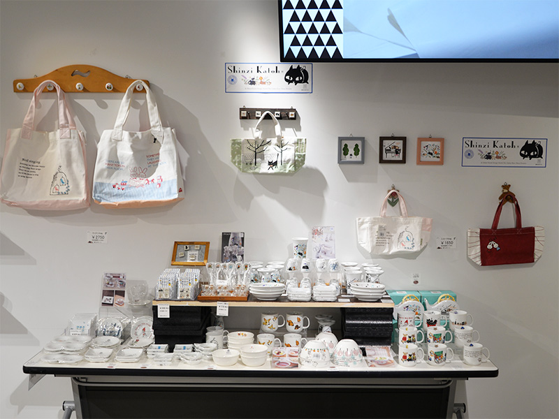 白い壁に木製のフックハンガーが取り付けられ、ピンクと白の猫のイラストが描かれたトートバッグが2つ掛けられている。右側の壁には、黒猫のイラストが描かれたブランドロゴや、猫と風景のイラストが飾られた額縁が並んでいる。下のテーブルには、猫や花柄の可愛らしいデザインの食器類が多数陳列されている。