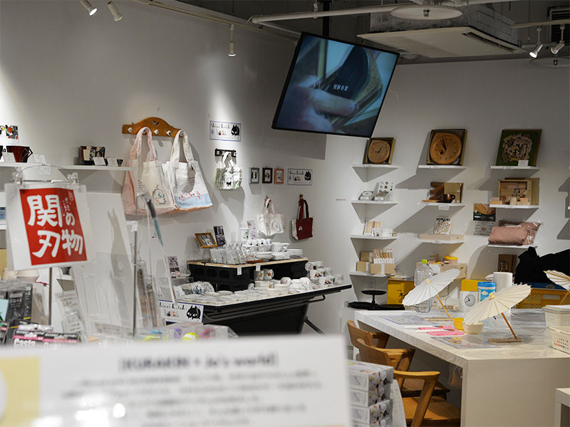 ギフトショップの店内風景。左側には「関の刃物」と書かれた赤い看板が見える。中央のテーブルには猫のイラストが入った白い食器類や雑貨が並び、奥の棚には木製の時計や雑貨などがディスプレイされている。天井にはモニターが設置されている。