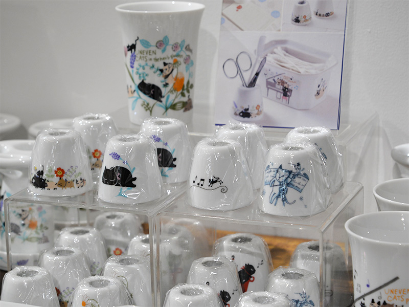 透明なプラスチックケースに陳列された、様々な猫のイラストが描かれた白い陶器製のタンブラーやコップ。それぞれのコップには異なるポーズやデザインの黒猫が描かれており、背景には音符や花柄が見えるものもある。