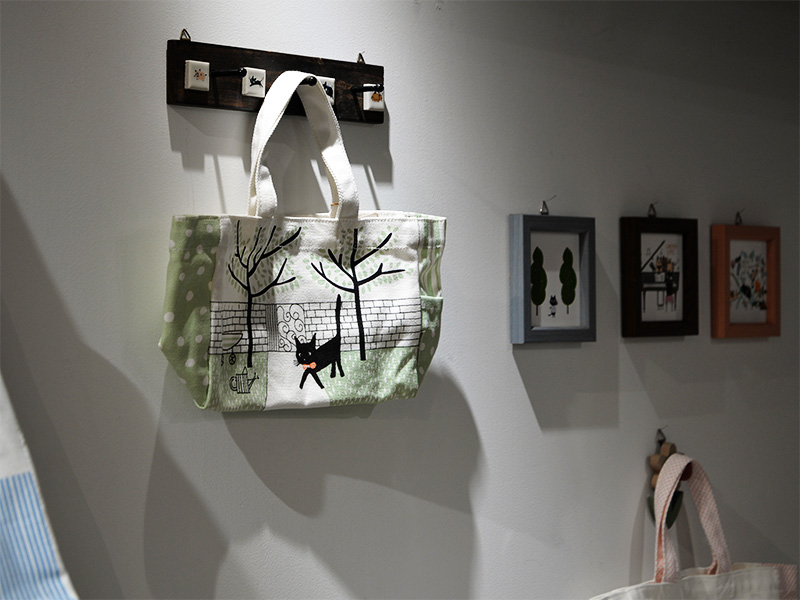 白い壁に取り付けられた木製のフックに、緑と白のチェック柄と猫のイラストが組み合わされた布製のバッグが掛けられている。壁には猫のイラストや風景画の入った額縁がいくつか飾られている。