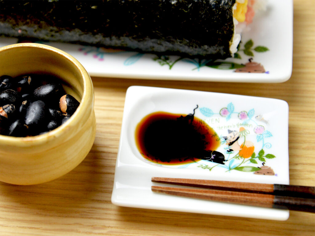 雑貨デザイナーシンジカトウが箸置きと小皿が一緒になった白い美濃焼製の箸置き小皿に可愛い猫のイラストが描かれ、この小皿にお醤油を入れてそばには巻きずしが置いてあるイメージ画像です。