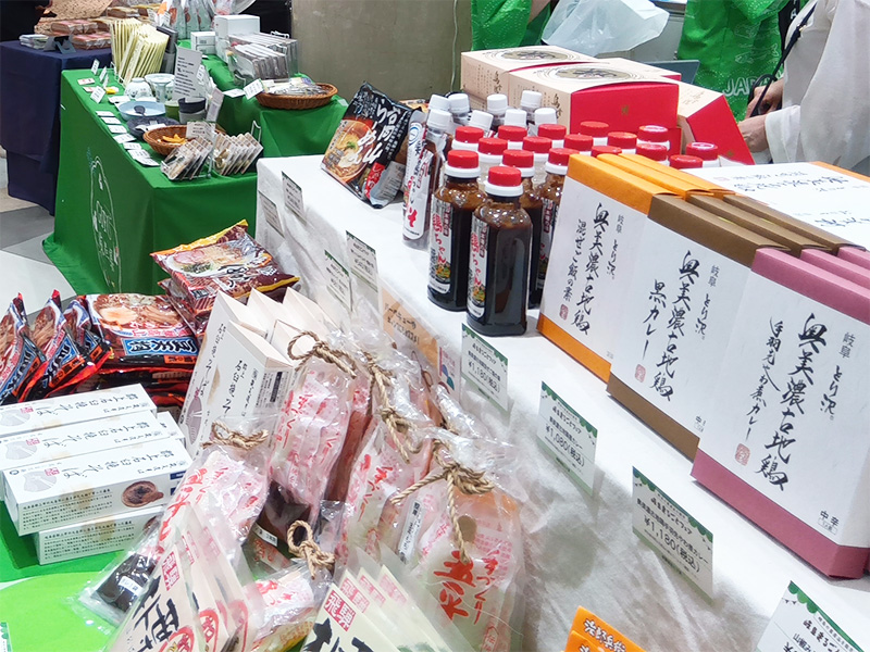 「岐阜まるごとフェア」の食品ブース。奥美濃古地鶏のカレーや五平餅のセット、そばなど、様々な岐阜の特産品が並べられています。手前には、袋に入った商品がいくつも積まれています。