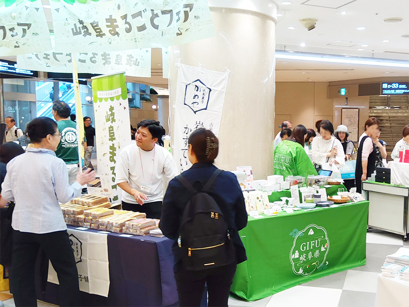 「岐阜まるごとフェア」の会場風景。緑のブースには、白いテーブルクロスがかけられ、「かめや」のカステラが箱に詰めて並べられています。ブースのスタッフが、来場者と笑顔で話しています。