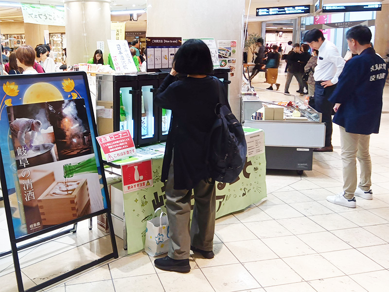  岐阜県の日本酒飲み比べコーナーの様子。黒い服を着た女性が、日本酒のサーバーをのぞき込んでいます。隣には、酒造りの様子が描かれた大きなパネルが立てられています。

