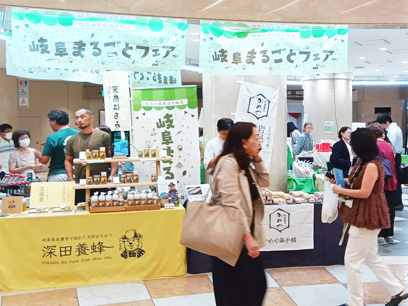 「岐阜まるごとフェア」の会場風景。上部には「岐阜まるごとフェア」と書かれた白い横断幕が吊るされています。ブースには、黄色いテーブルクロスをかけた「深田養蜂」のはちみつや関連商品が並び、多くの人が行き交っています。