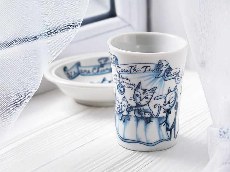 白い木製のテーブルの上に置かれたタンブラーと皿。窓から入る光に照らされ、透明な白いカーテンが優しく写り込んでいる。タンブラーの正面には、楽しげな猫たちのイラストがはっきりと見える。清潔感と温かみのある雰囲気が伝わる写真。