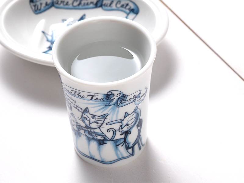 白い陶器製のタンブラー。表面には、青い線で描かれた愛らしい猫のイラストと文字がデザインされている。タンブラーには透明な水が約120ml入っており、手前には同じデザインの皿がぼんやりと写っている。清潔感のあるインテリアに馴染む、癒しの猫グッズ。