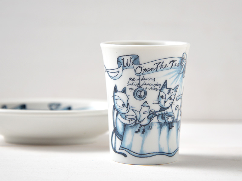 代替テキスト: 白い背景の前に置かれたタンブラーの正面写真。タンブラーには、雑貨デザイナーであるシンジカトウ氏による、ユーモラスなデザインの猫と文字のイラストが鮮明に描かれている。猫好きにはたまらない、日本製陶磁器のタンブラー。