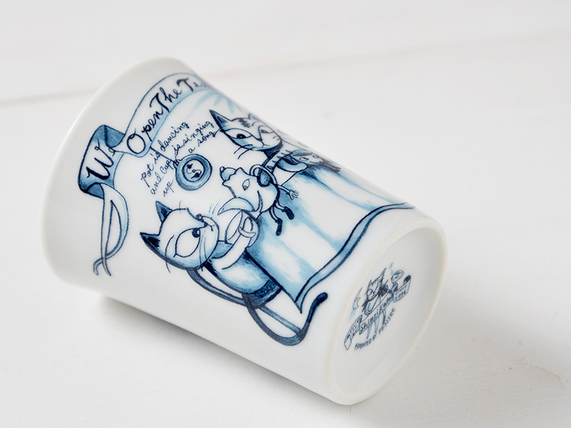 白い背景に、底面を斜めから写したタンブラーの写真。底面にはデザイナーの名前と製造元を示すロゴがはっきりと見える。全体に猫と文字のイラストが描かれ、デザイン性の高さが伝わる。日本製陶磁器の品質の良さが際立つカット。