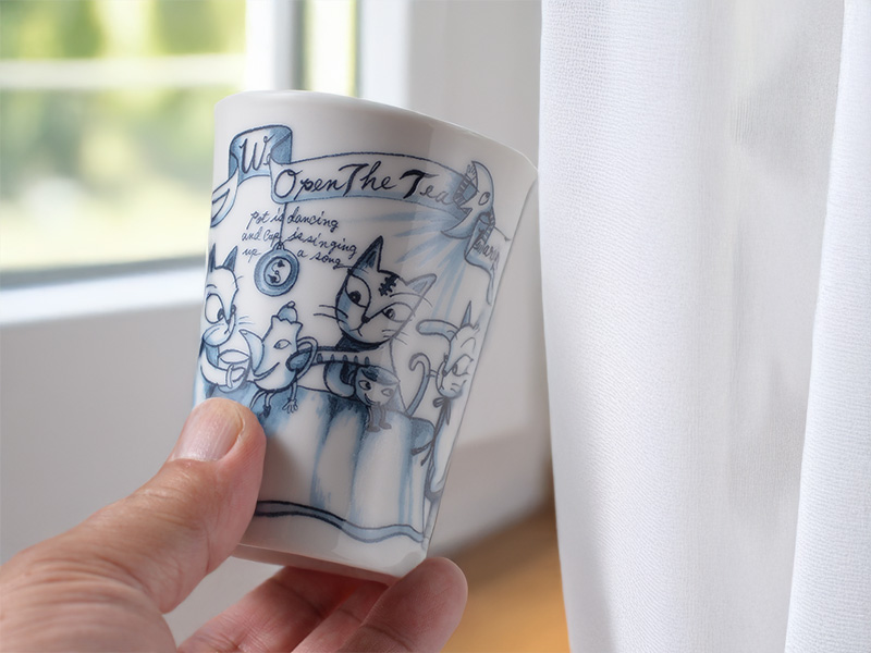 窓から柔らかな光が差し込む部屋で、男性が片手でタンブラーを持っている写真。タンブラーには、楽しげな猫たちのイラストが描かれている。白く清潔感のあるカーテンが背景に写っており、リラックスした雰囲気を伝えている。