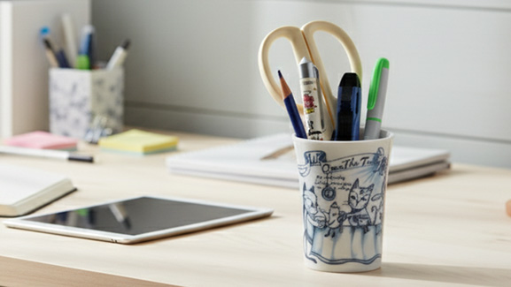 木製の机の上に置かれた白いタンブラー。タンブラーには色鉛筆やハサミ、ペンなどの文房具が入っており、ペン立てとして使用されている。周りにはタブレットやノート、ファイルが配置され、リモートワークや在宅勤務のデスク周りをおしゃれに整理している様子。