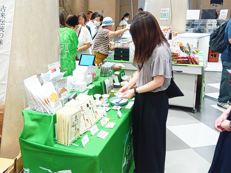  大阪のディアモール大阪で開催中の「岐阜まるごとフェア」の様子。多くの来場者が賑わう会場で、緑のテーブルクロスがかけられたブースに立つ女性が、陳列された商品を熱心に見ています。