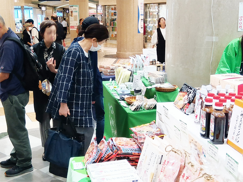 「岐阜まるごとフェア」のゼルポティエブース。チェック柄のシャツを着た女性が、猫のイラストが描かれた美濃焼の茶碗や、青い小皿、白い箸置きなど、可愛らしい陶器の商品を手に取って見ています。ブースには様々な商品が並び、多くの来場者が賑わっています。