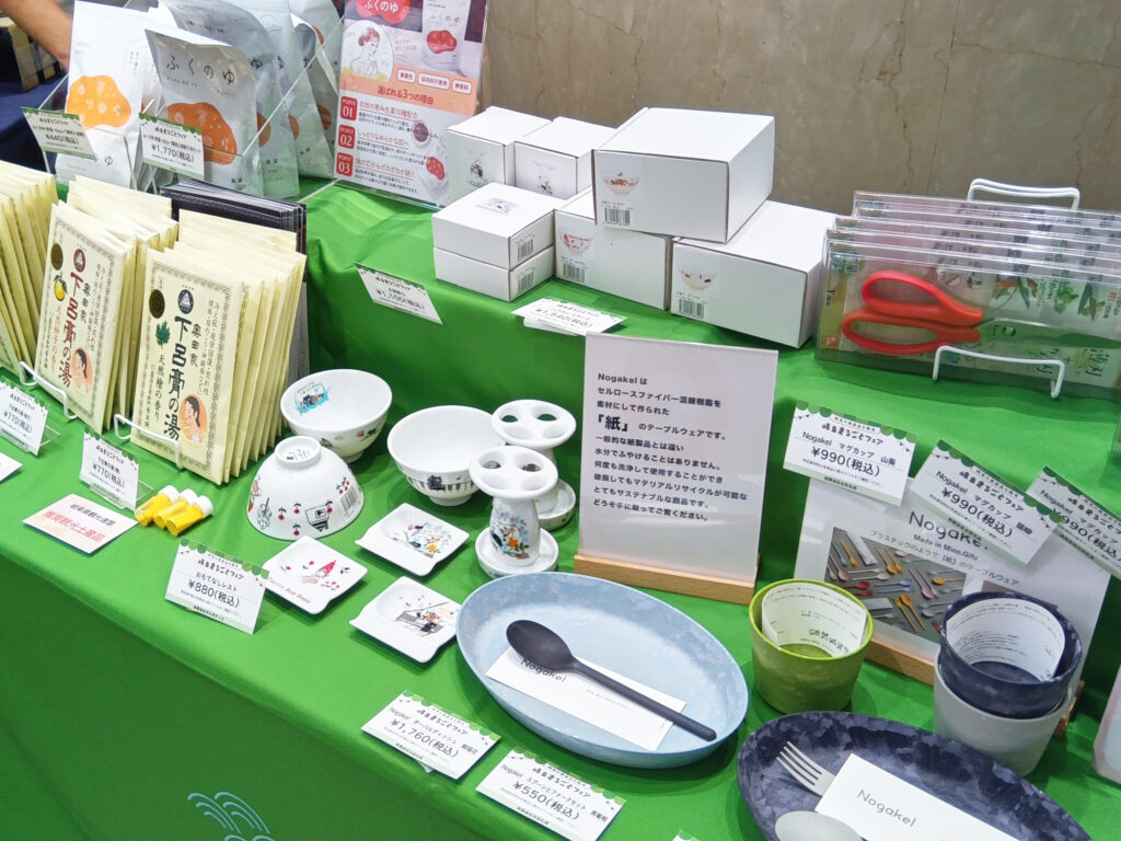 ゼルポティエの美濃焼商品。白いテーブルに、かわいい猫や赤ずきんちゃんのイラストが描かれた茶碗や箸置き、歯ブラシスタンドなどが並べられています。隣には、美濃和紙で作られたお皿や、下呂の入浴剤も陳列されています。