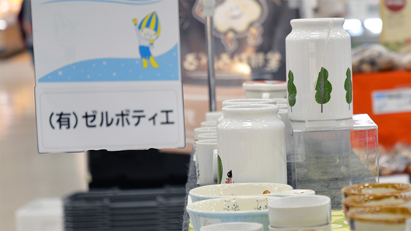 「(有)ゼルポティエ」と書かれた白い看板が手前にあり、その奥には、可愛らしい木のイラストや猫の模様が描かれた白い陶器の保存容器や食器が並べられた売り場の様子。右奥には別の催事の様子もぼんやりと見える。
