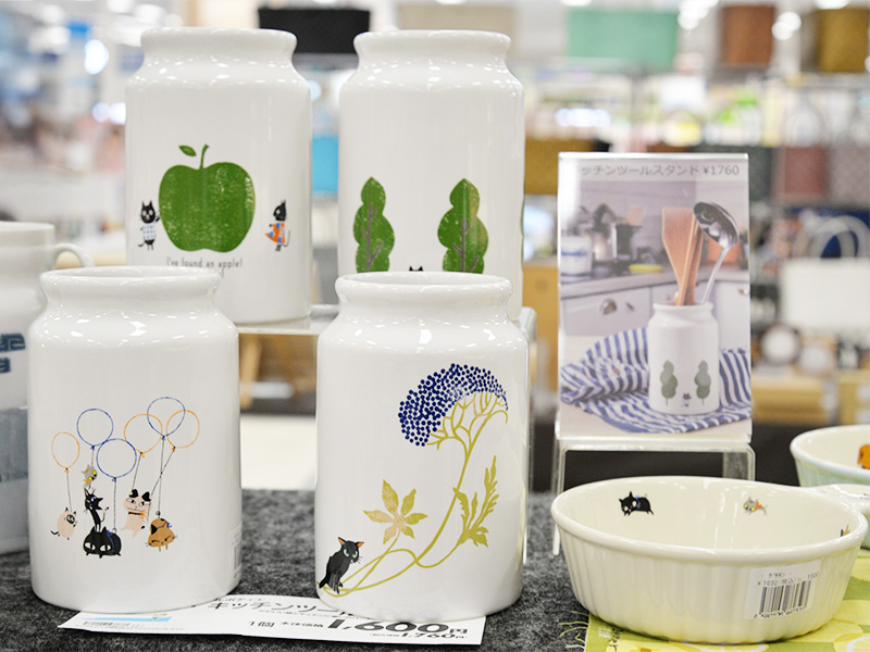 猫やリンゴ、花などのイラストが描かれた、白い陶器製のキッチンツールスタンドや保存容器が4つ並んでいる。