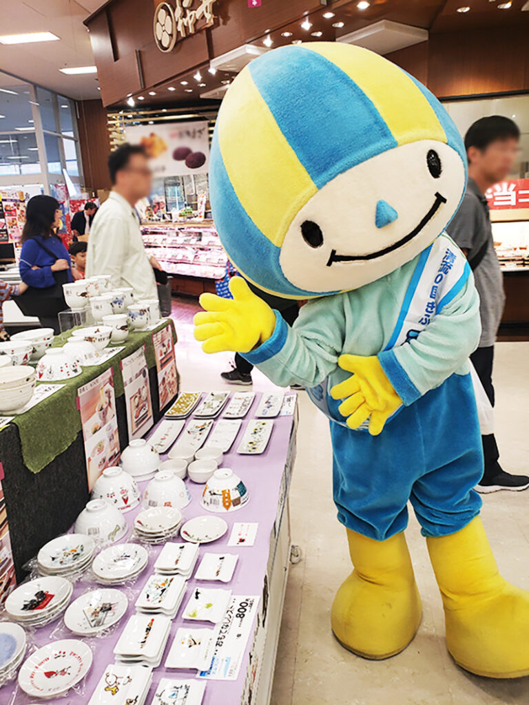 岐阜県のゆるキャラ「ミナモ」が、ゼルポティエの食器が並ぶブースの前で、笑顔で手を広げている様子。周囲には買い物客の姿が見える。