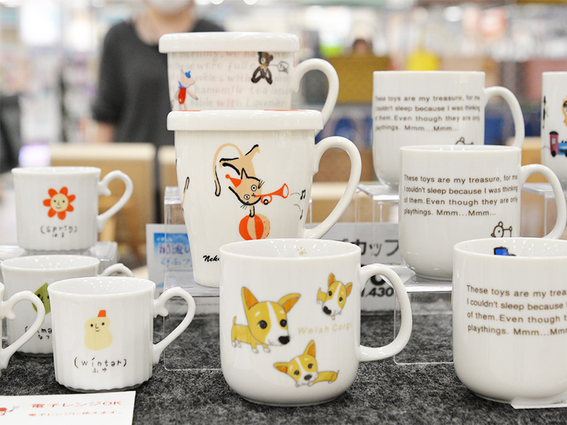 ウェルシュ・コーギーや、蓋付きでラッパを吹く猫のイラストが描かれた、可愛らしいデザインの白いマグカップが多数陳列されている。