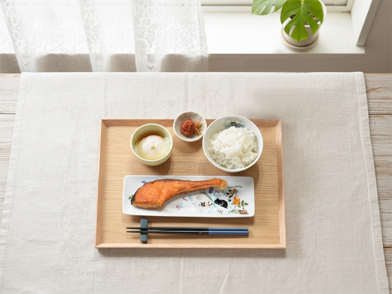 猫のイラストが描かれた横長の皿に焼き魚を、小皿にご飯と温泉卵を添えた、和風の朝食の食卓風景。猫のイラストがデザインされた食器を使った和風の食卓。猫好き必見の可愛い猫柄プレート。