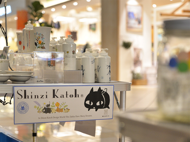 【ブランドロゴ】催事テーブル手前に掲げられた、シンジカトウ氏の黒猫キャラクターのブランドロゴ。その後ろには、猫のイラストが描かれたディスペンサーなどの雑貨が並んでいる。