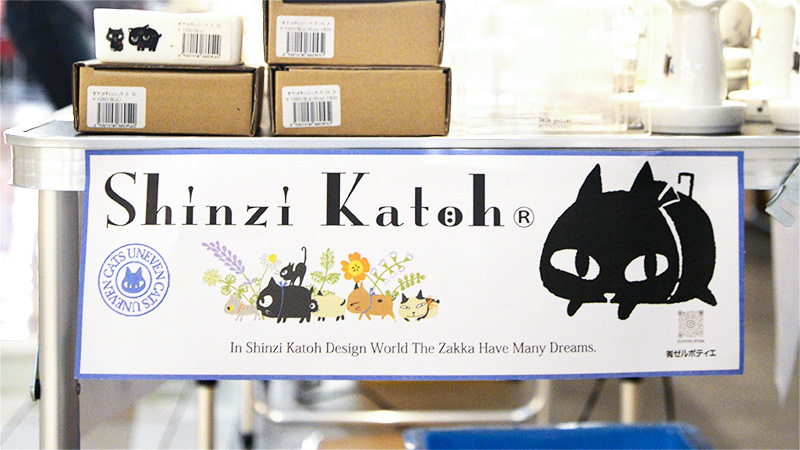 ゼルポティエ催事会場に設置された、シンジカトウ氏のブランドロゴ看板。大きな黒猫のキャラクターと、可愛い動物たちが草花の中で遊ぶイラストが描かれ、「In Shinzi Katoh Design World The Zakka Have Many Dreams.」というメッセージが添えられている。ブランドの世界観を象徴するデザイン。