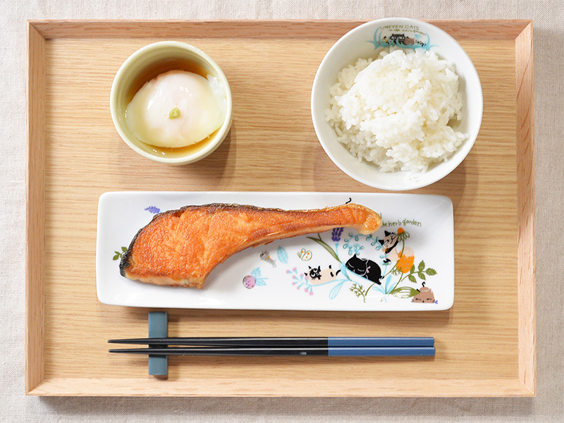 【商品使用例】シンジカトウデザインの長皿に盛り付けられた焼き鮭と、ご飯、温泉卵などの和朝食。器が日々の食卓を優しく彩っている様子。