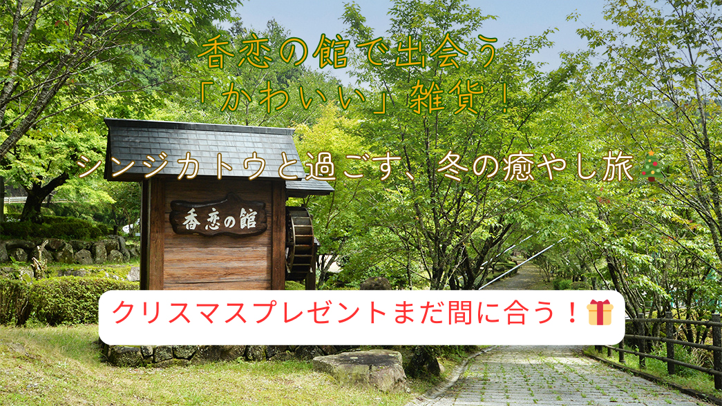緑豊かな木々に囲まれた「香恋の館」の入り口と、そこへ続く石畳の道を背景にしたブログアイキャッチ画像。画面中央には、小さな水車が付いた趣のある木製の看板が立っています。画像上部には黄色い文字で「香恋の館で出会う『かわいい』雑貨！」、中央には白い文字で「シンジカトウと過ごす、冬の癒やし旅」というタイトルが重なっています。下部には、赤いリボンが付いたプレゼントボックスの絵文字と共に、白い背景に赤文字で「クリスマスプレゼントまだ間に合う！」というキャッチコピーが強調されています。