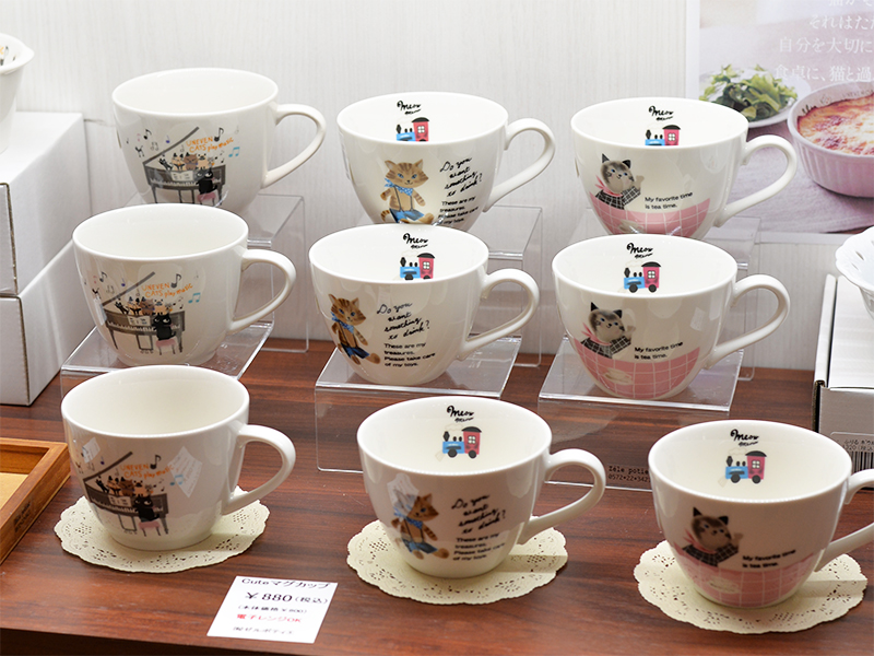 丸猫展の棚に整列した白いマグカップ。ピアノを弾く猫や、汽車に乗る猫など、シンジカトウ氏らしいユーモアあふれるイラストが描かれています。カップの内側にも小さな猫が隠れていて、使うたびに目が合う楽しさがあります。