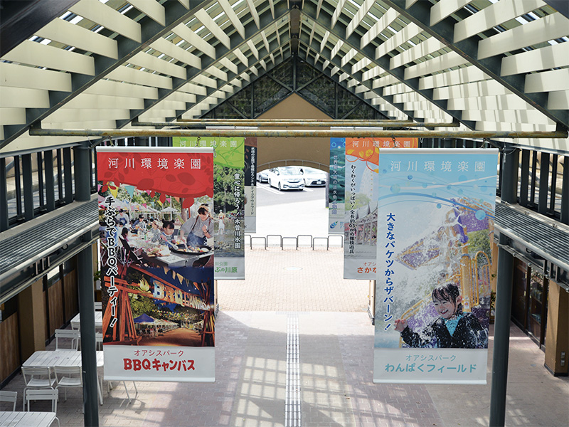 オアシスパーク園内の通路。黒い鉄骨と白いルーバーで構成された高い屋根の下に、「河川環境楽園」「BBQキャンパス」「わんぱくフィールド」などのイベントや施設を紹介する色鮮やかな縦長のバナー（垂れ幕）が複数掛けられている。