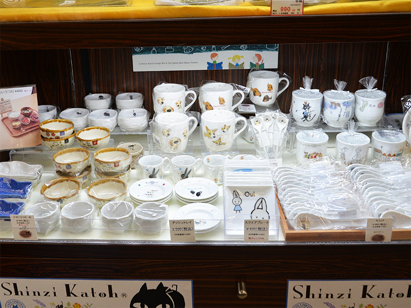 「岐阜おみやげ川島店」のゼルポティエ（zelepotier）コーナー。Shinzi Katoh（シンジカトウ）デザインのマグカップ、小鉢、皿、箸置きなどが木製棚にきれいに並べられている。上部には「Shinzi Katoh Design」のロゴパネル。
