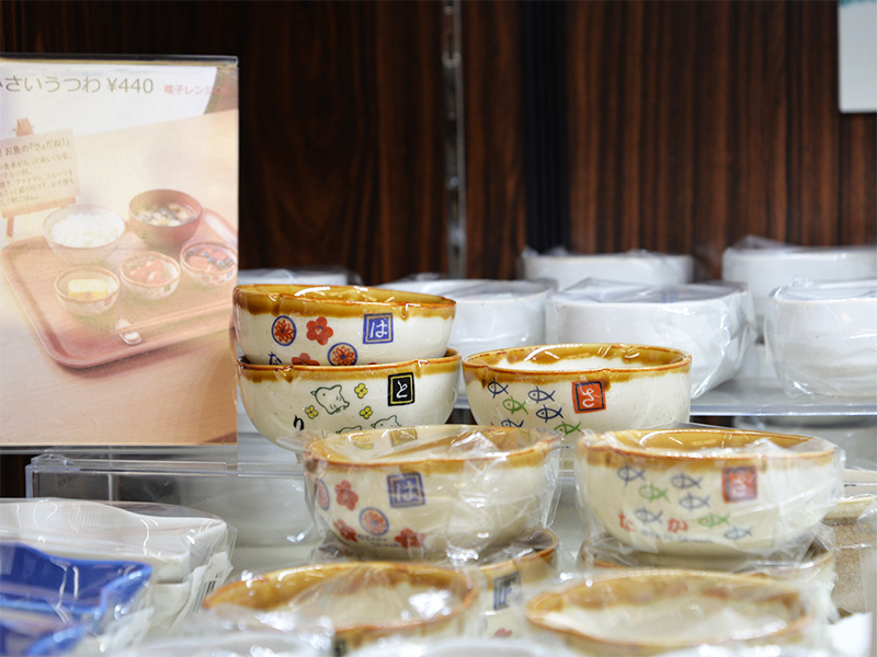 「ちいさい器」として陳列された、子ども向けの和食器。白い小鉢の側面には、ひらがなや、魚、梅の花、鳥などの可愛いイラストと、「Designed by Shinzi Katoh（シンジカトウ）」の文字がデザインされている。