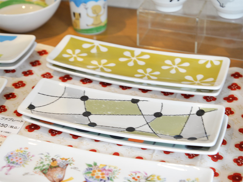 【たじみ陶器まつり】シンジカトウデザインの人気シリーズ「アルファプレート」と長皿の展示。スタッキング（積み重ね）された使い勝手の良いサイズの平皿が並び、表面には小鳥や植物、英字などの繊細なイラストが描かれている。シンプルながら温かみのある美濃焼の器が、木製の棚に整然とディスプレイされている様子。