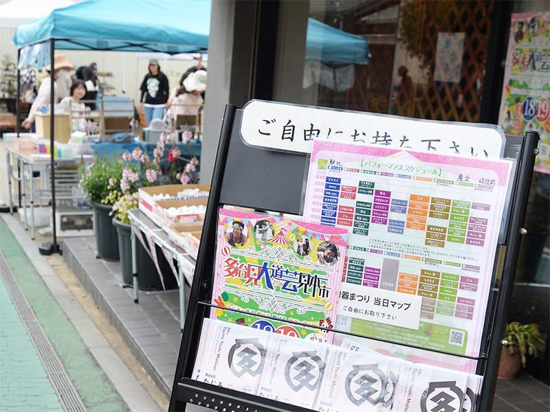 【たじみ陶器まつり】呉服屋「みかわや」の店先に置かれた、イベント案内用のパンフレットラック。第84回たじみ陶器まつりのチラシが並び、その奥にはシンジカトウデザインのモダンな角皿がディスプレイされている。お祭りの情報を求めて人々が立ち寄る、活気ある店先風景。