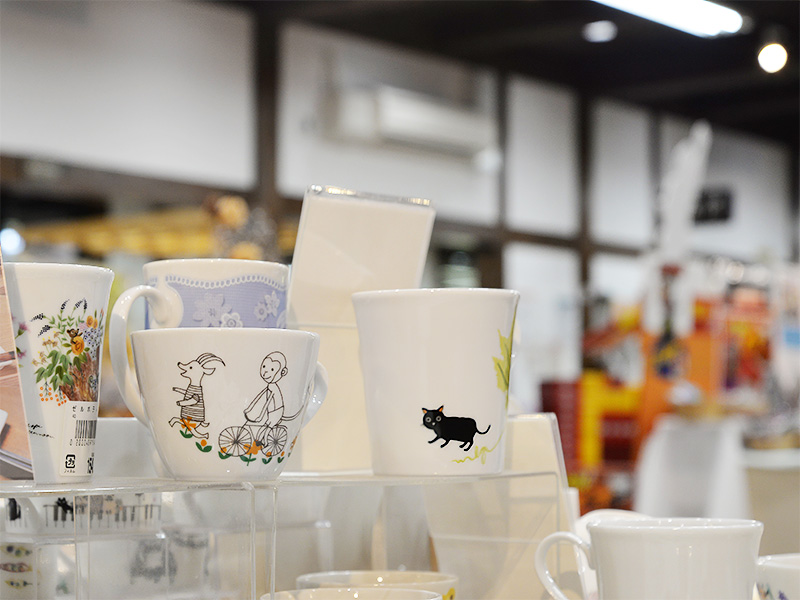 10月2日に「香恋の館」へ商品補充を行った際の展示棚。黒猫が歩くデザインや、自転車に乗るサルとヤギの楽しげなイラストなど、Designed by Shinzi Katoh(シンジカトウ)のマグカップやフリーカップが透明なアクリルスタンドの上に可愛らしく並べられています。