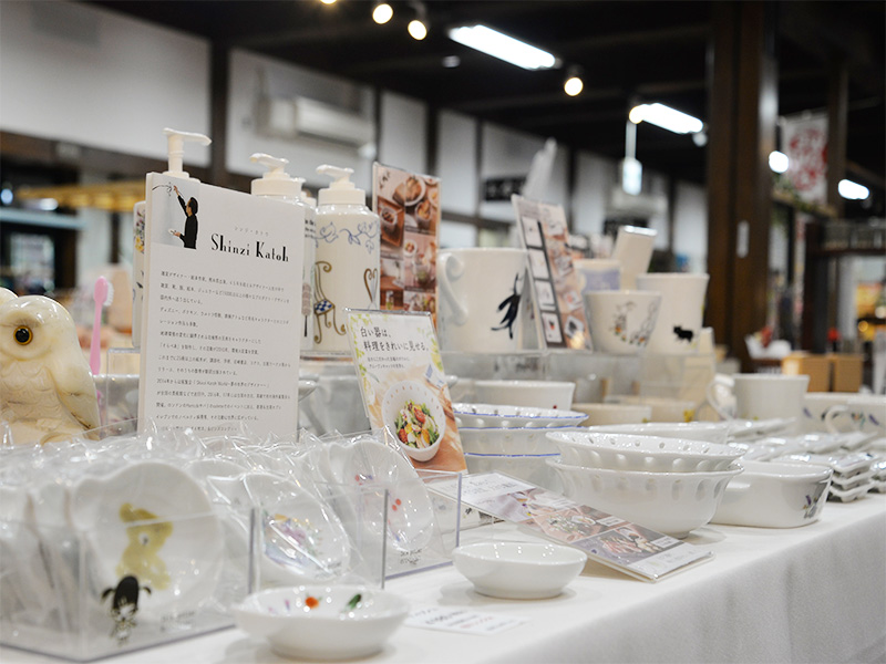 10月2日に「香恋の館」へ商品補充を行った際の展示スペース。「Shinzi Katoh」の紹介POPを中心に、Designed by Shinzi Katoh(シンジカトウ)の透かし彫り入りの白いボウルや小皿、マグカップ、ディスペンサーなどの陶器商品がテーブルいっぱいに美しくディスプレイされています。