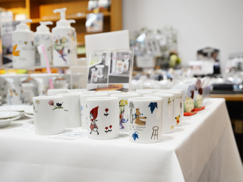 10月2日に「香恋の館」へ商品補充を行った際の展示コーナー。白いテーブルクロスの上に、赤ずきんやクマ、黒猫などDesigned by Shinzi Katoh(シンジカトウ)の可愛いイラストが描かれたフリーカップやタンブラーがずらりと美しく並べられ、奥には陶器製のソープディスペンサーも陳列されています。