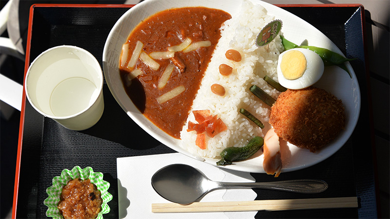 黒いお盆の上に載った香恋の館の名物カレー。ダムに見立てた山盛りの白いご飯の片側に濃厚なカレーソースがかかり、ゆで卵、コロッケ、ウインナー、漬物、福神漬け、とろけるチーズが彩り豊かに添えられています。