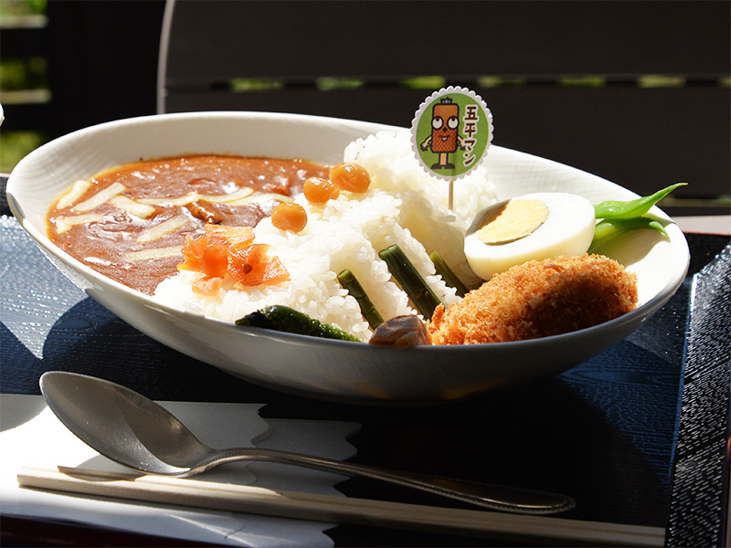 10月2日に「香恋の館」へ商品補充に訪れた際のお昼ご飯。白い大きなオーバル深皿に盛り付けられた名物の羽布ダムカレー。ダム堤体に見立てた白いご飯の上に「五平マン」のかわいいキャラクターピックが挿され、カレーソース、サクサクのコロッケ、ゆで卵、インゲン、福神漬けなどが添えられています。